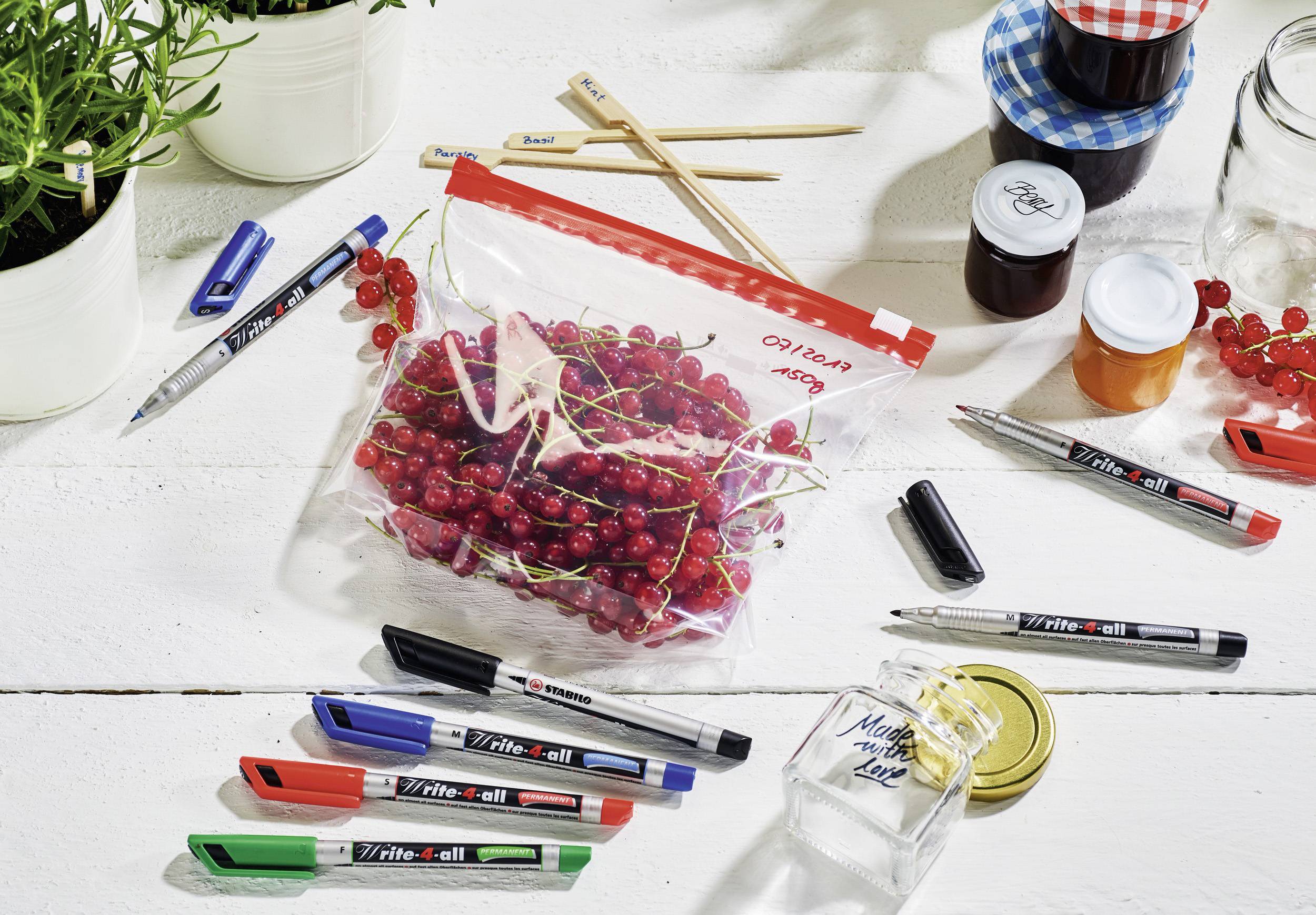 Plastic bag of red currants on a table, surrounded by pens, glasses and plants.
