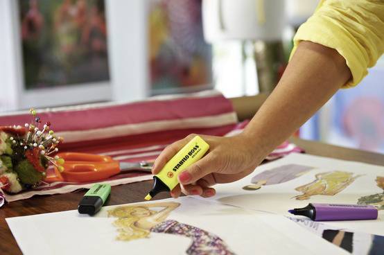 A person is sketching fashion designs on paper using coloured highlighters. Fabrics and sewing accessories are scattered in the background.