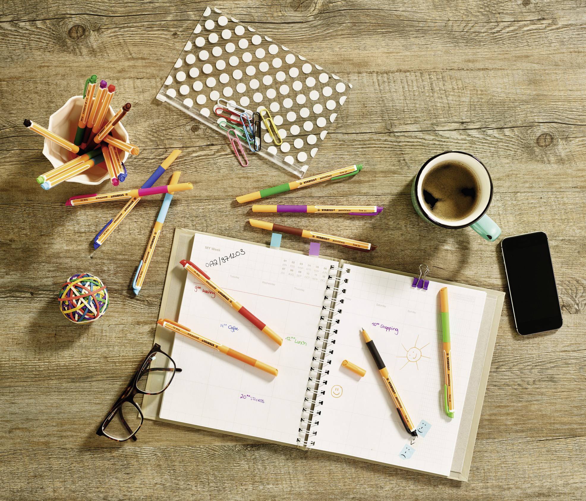 An open planner with colourful pens lies on a wooden table next to a pair of glasses, a mobile phone, a coffee cup, and a pencil pot.