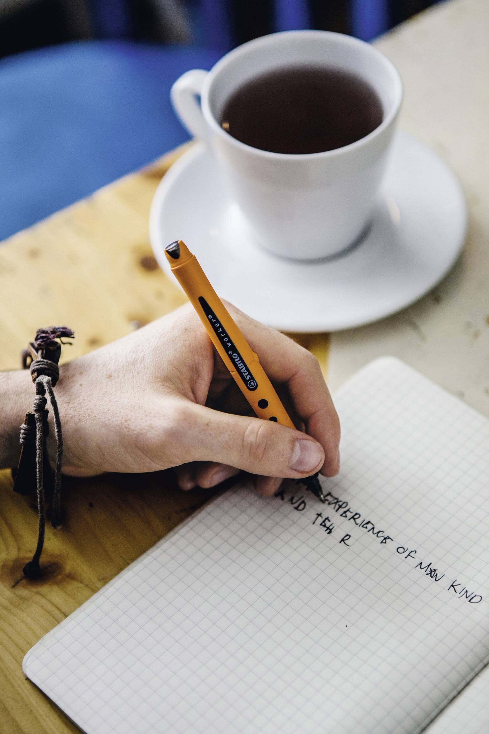 A person is writing in a notebook on a table. Next to it sits a cup of coffee.