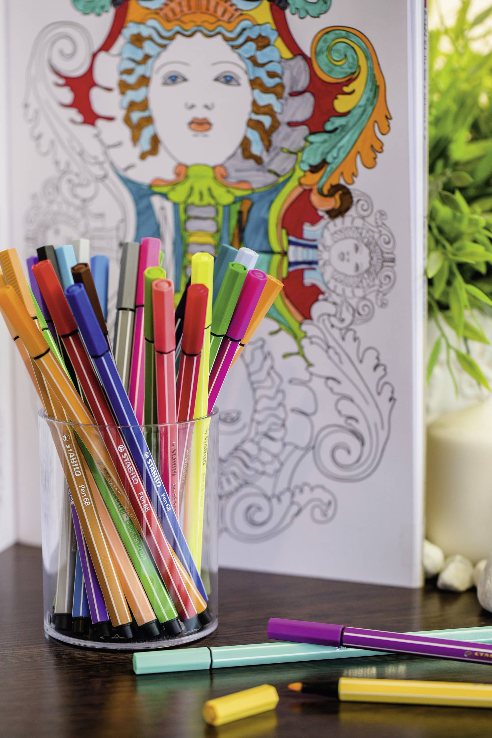 A glass filled with colourful felt-tip pens sits in front of a partially coloured colouring book with a decorative facial pattern.