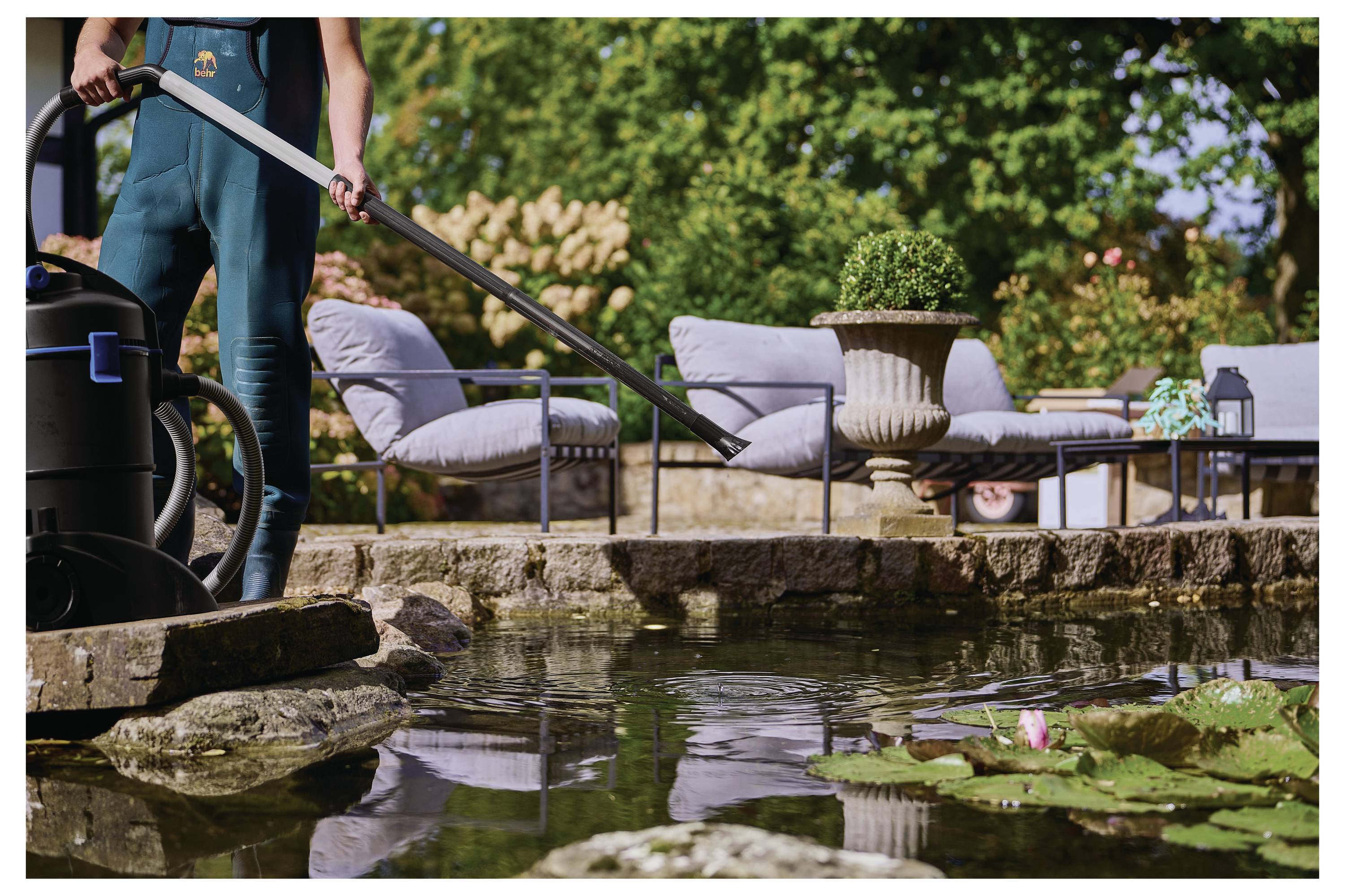 Person cleaning a pond with a vacuum cleaner; stone edge, water lilies, outdoor seating, and plants in background suggest a garden setting.
