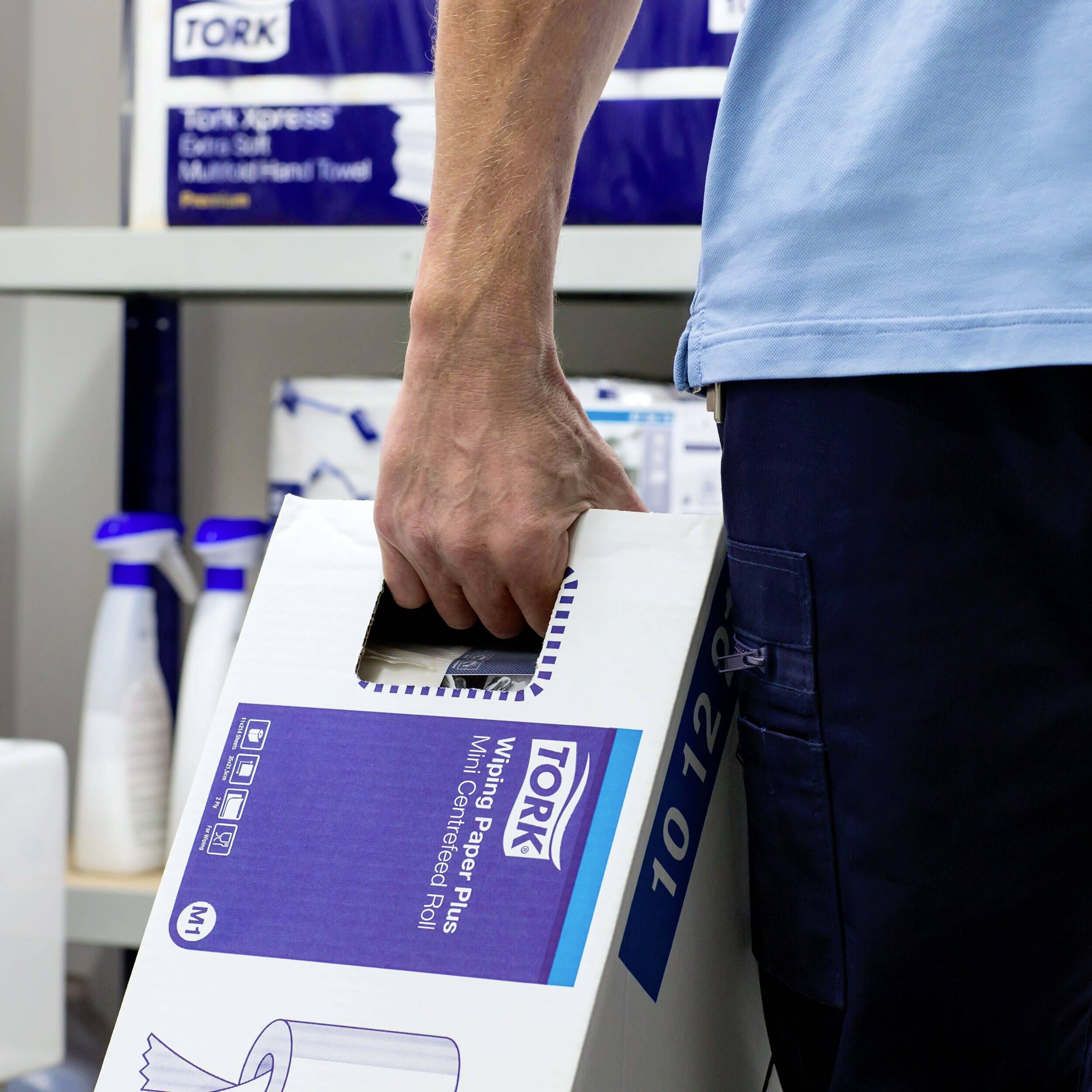 A person is carrying a box of paper towels in a storage room. In the background, there are additional cleaning products and shelves.