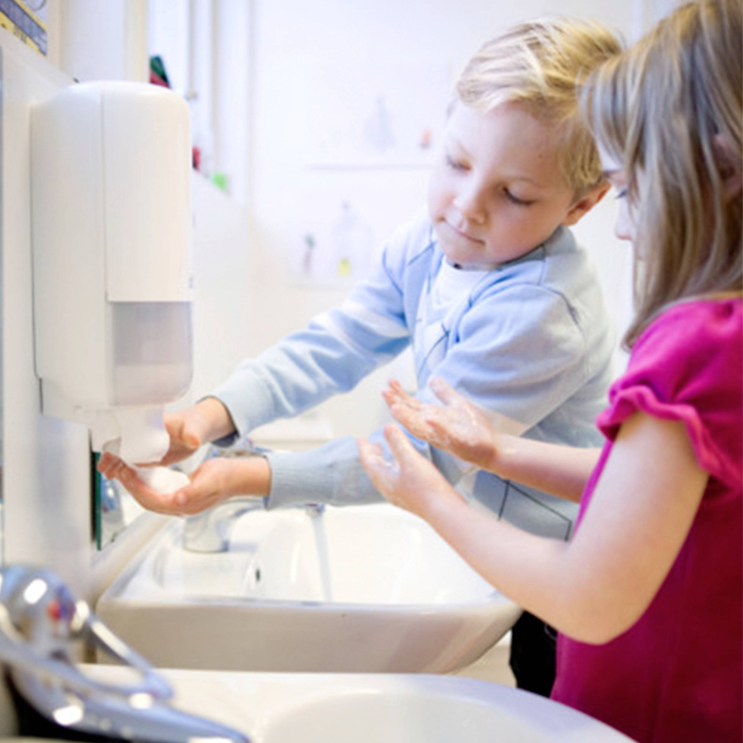 A boy and a girl are washing their hands at a sink. The boy is squeezing soap from a dispenser.
