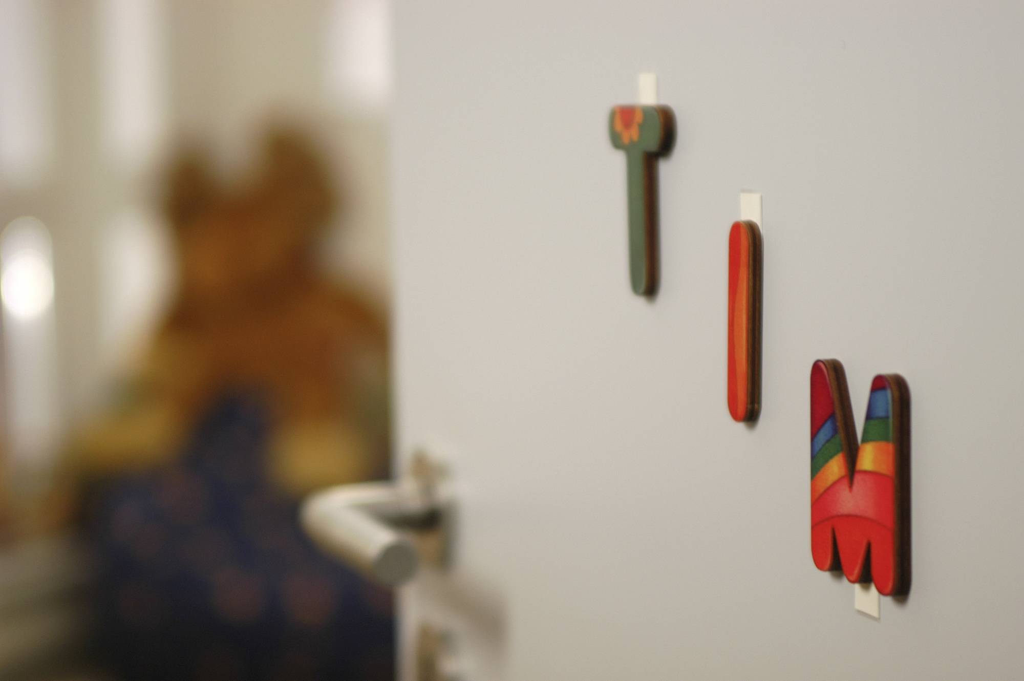 Door with coloured wooden letters 'T I M'; blurred background shows hint of a child's bedroom with soft toy.