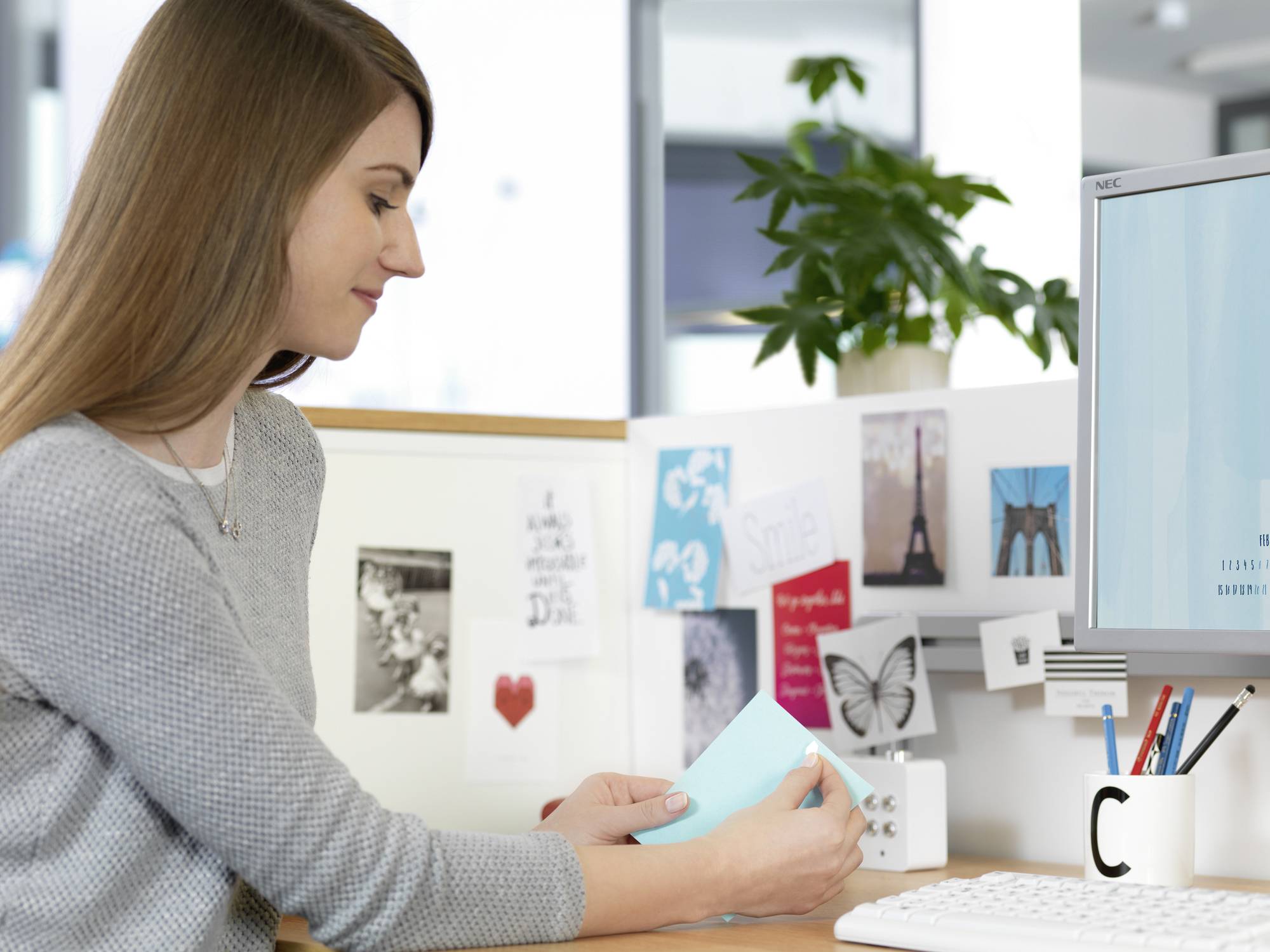 A woman is sitting at a desk reading a document. In the background, there are photographs, maps, and a plant. Desk with a computer monitor and a mug.