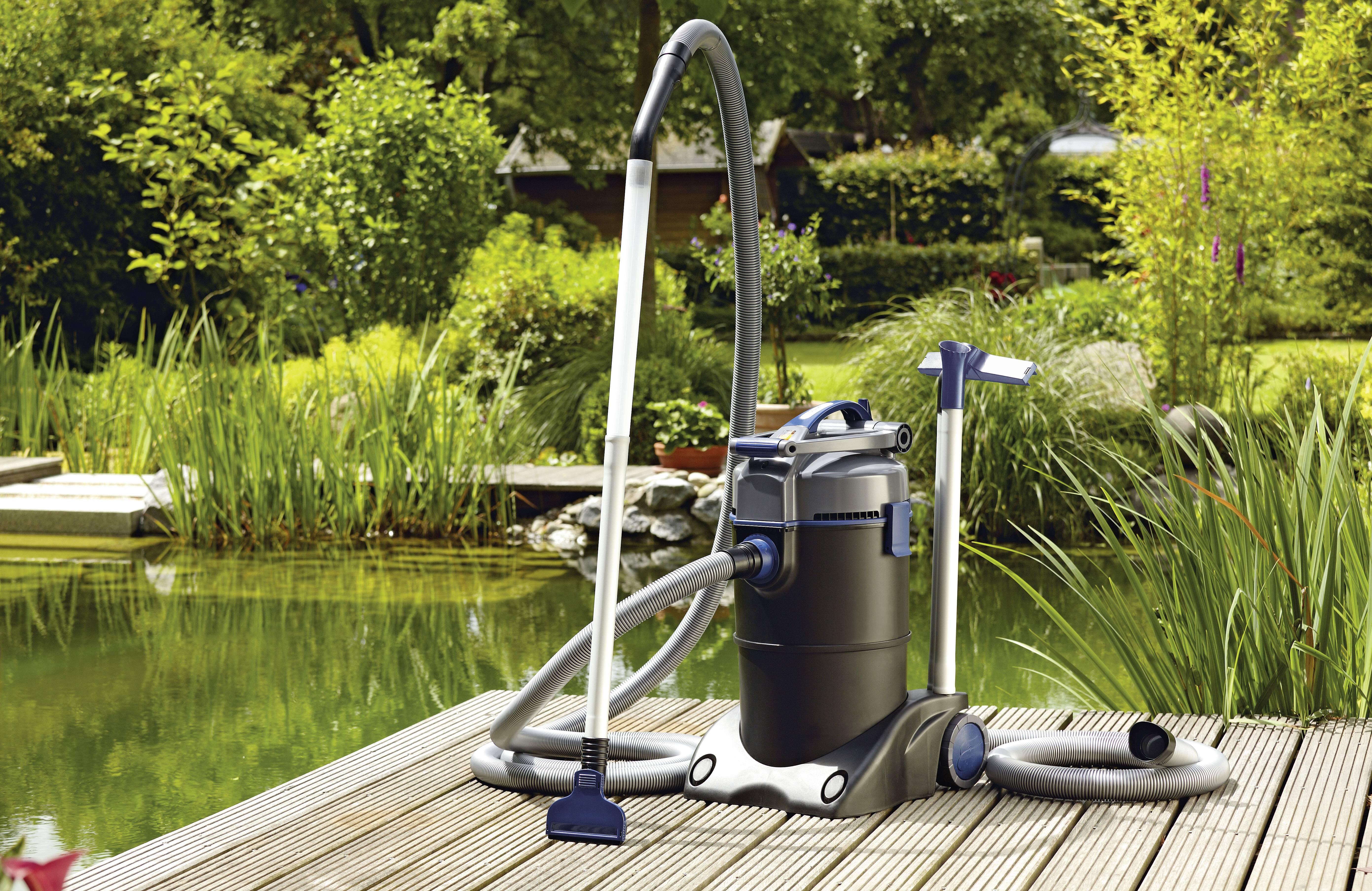 A wet/dry vacuum cleaner with hoses and attachments sits on a wooden deck near a pond surrounded by lush greenery.