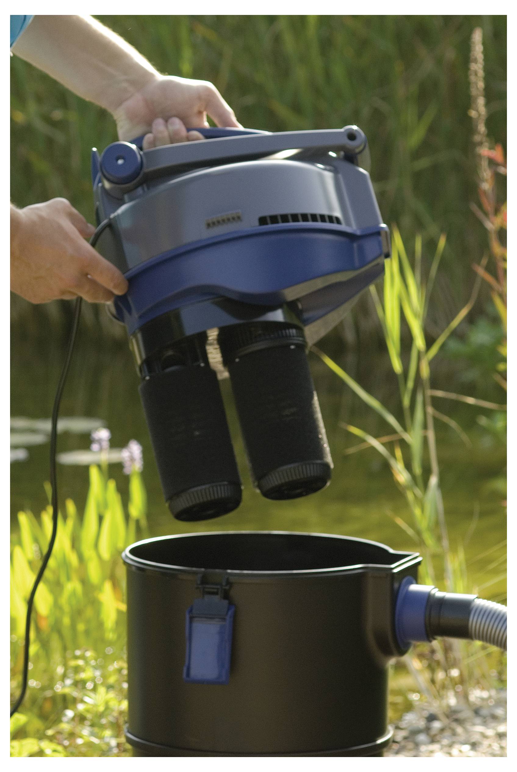 A person holds a blue and gray vacuum cleaner above a black container in an outdoor setting with grass and water in the background.