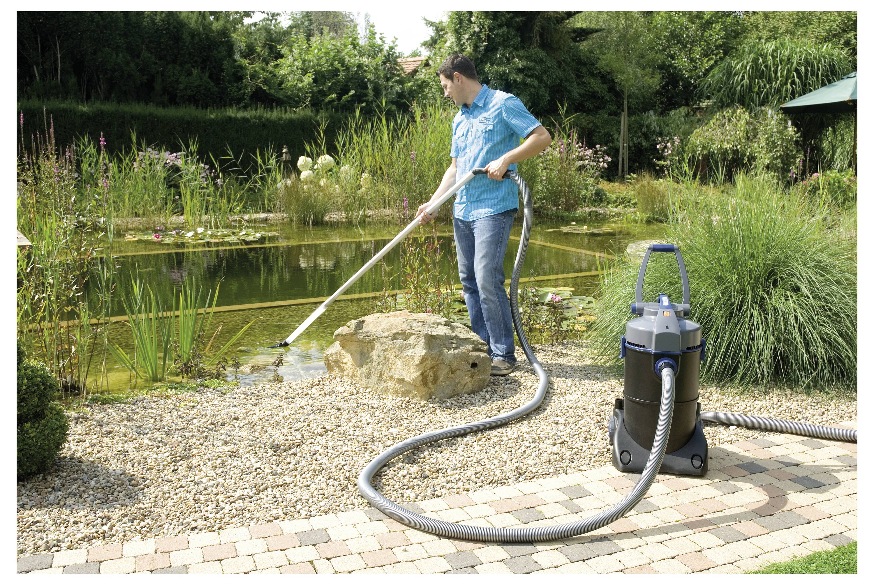 Man using a vacuum-like device to clean a pond near a garden area, with water plants and neatly arranged rocks in the background.