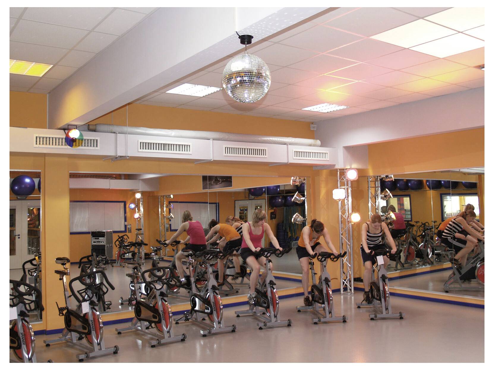 People exercise on stationary bikes in a mirrored gym studio with bright lights and a disco ball.