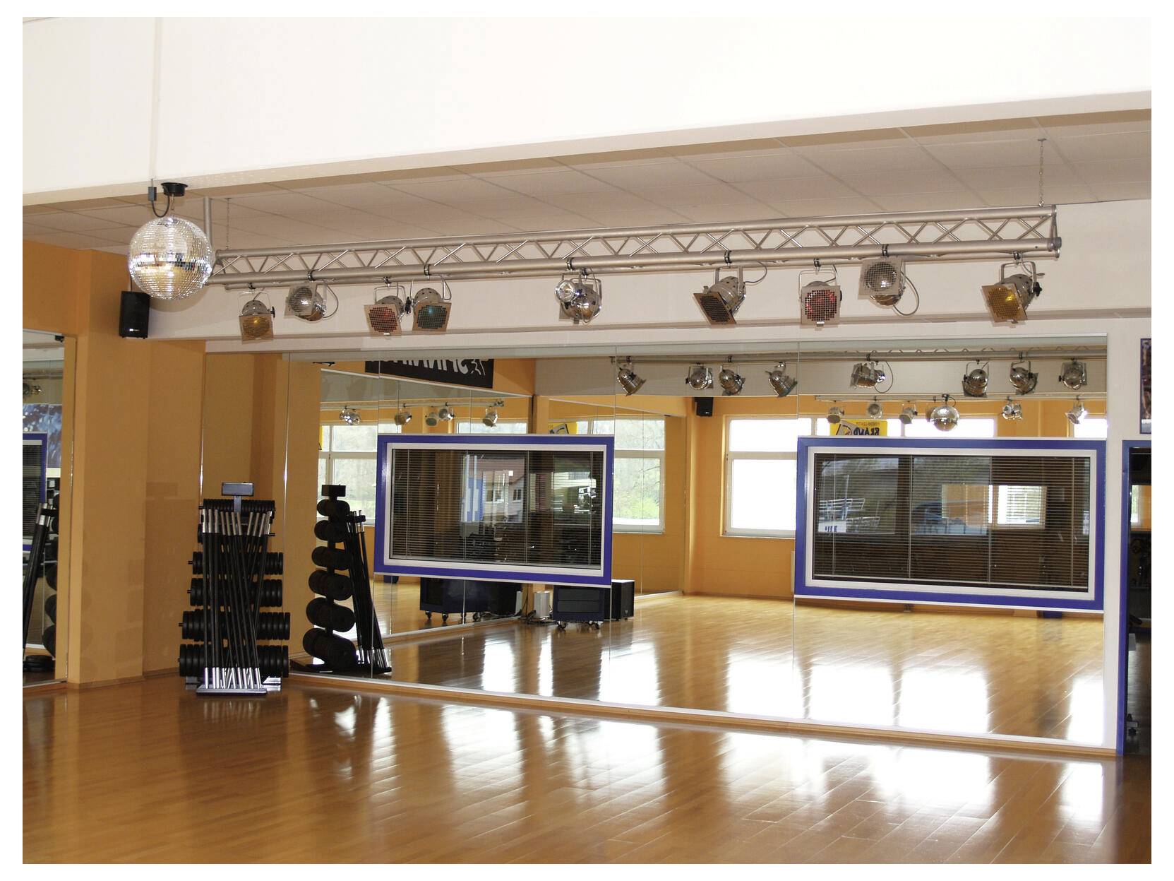 Empty dance studio with a mirrored wall, wooden floor, and ceiling-mounted stage lights; stacked step platforms sit at the left.
