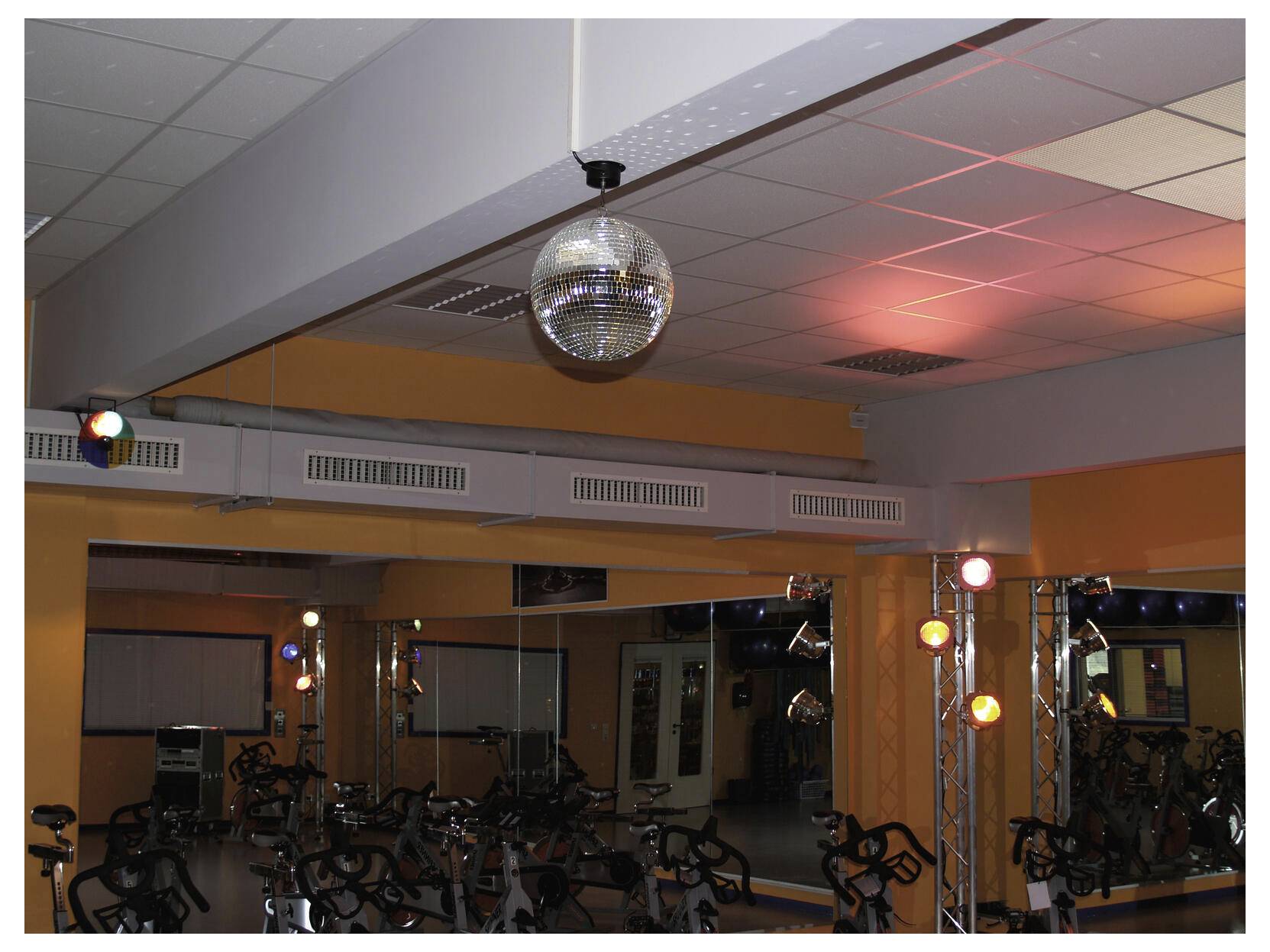 A mirrored disco ball hangs in a gym or studio with exercise bikes, lit by colored stage lights reflecting on the ceiling and walls.