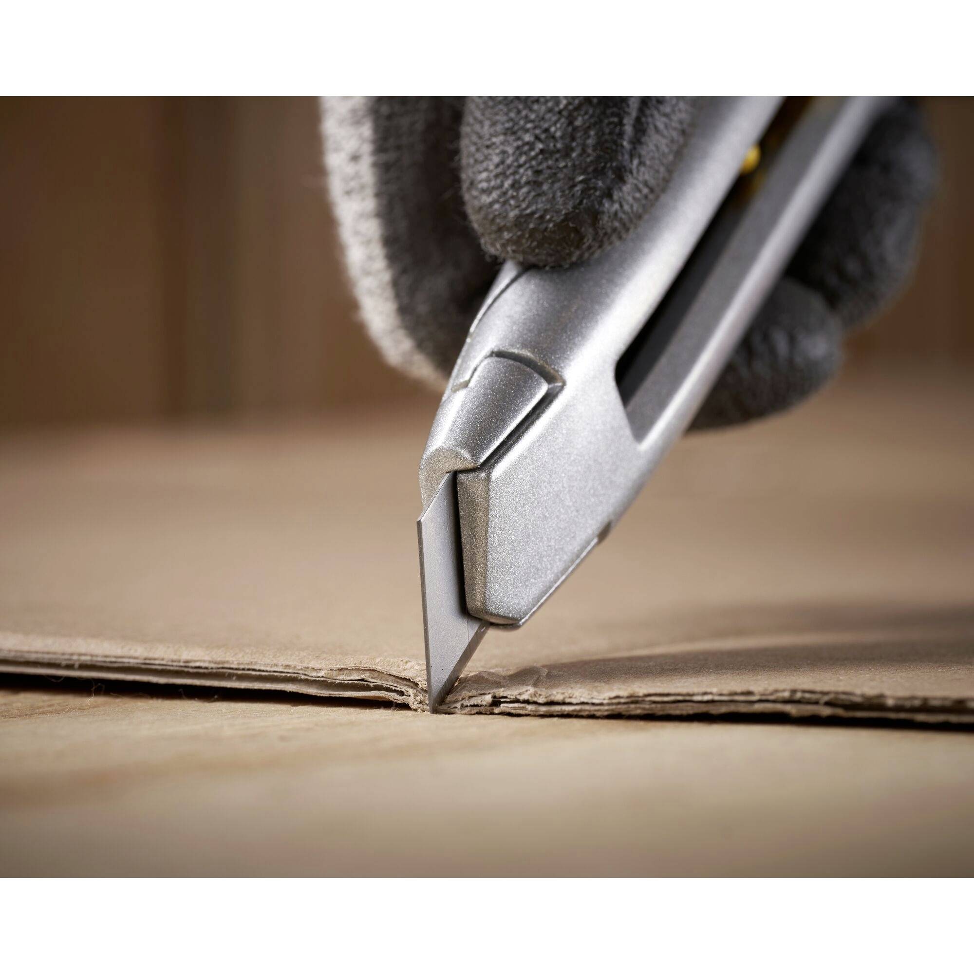 A gloved hand uses a silver craft knife to cut a cardboard box precisely.