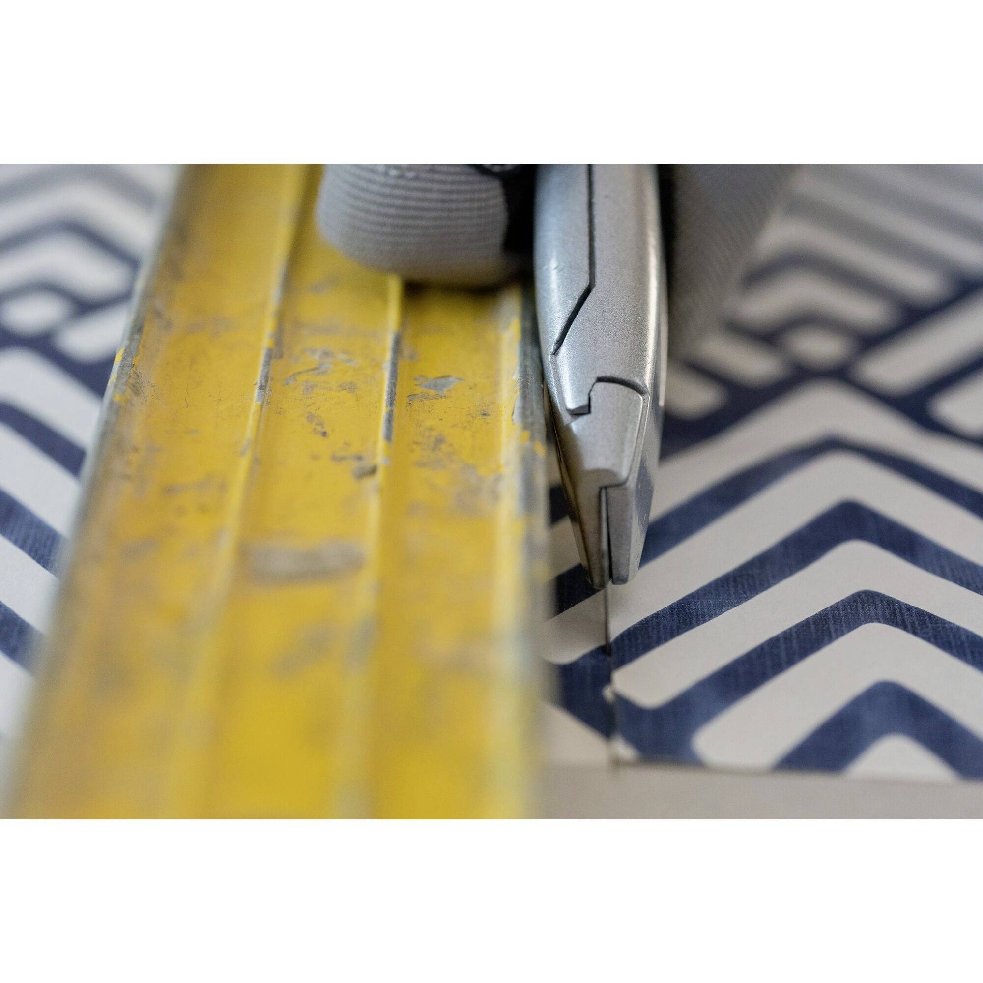 A hand wearing a glove is cutting a chevron pattern on paper with a craft knife along a yellow ruler.