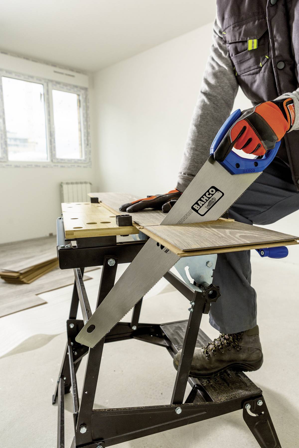 Eine Person sägt Holz mit einer Handsäge auf einer Arbeitsbank in einem renovierten Raum. Sie trägt Arbeitshandschuhe.