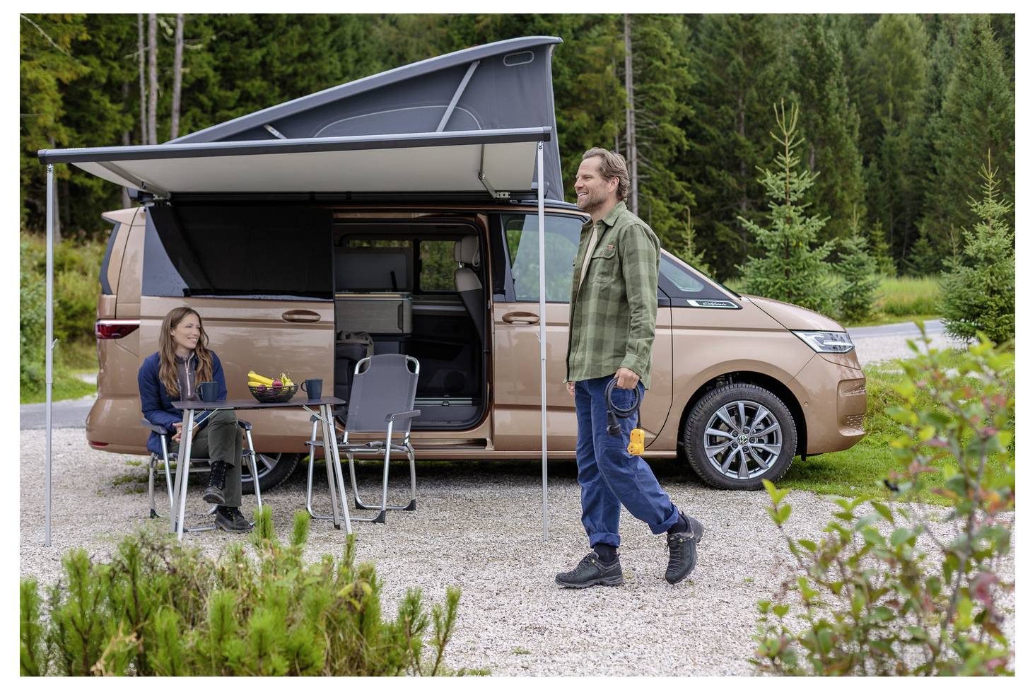 Two people camping beside a tan camper van with pop-up awning; one sits at a small table while the other walks nearby in a forest clearing.