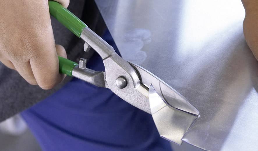 A person is cutting a piece of metal sheet with a tin snips. The snips have green handles.