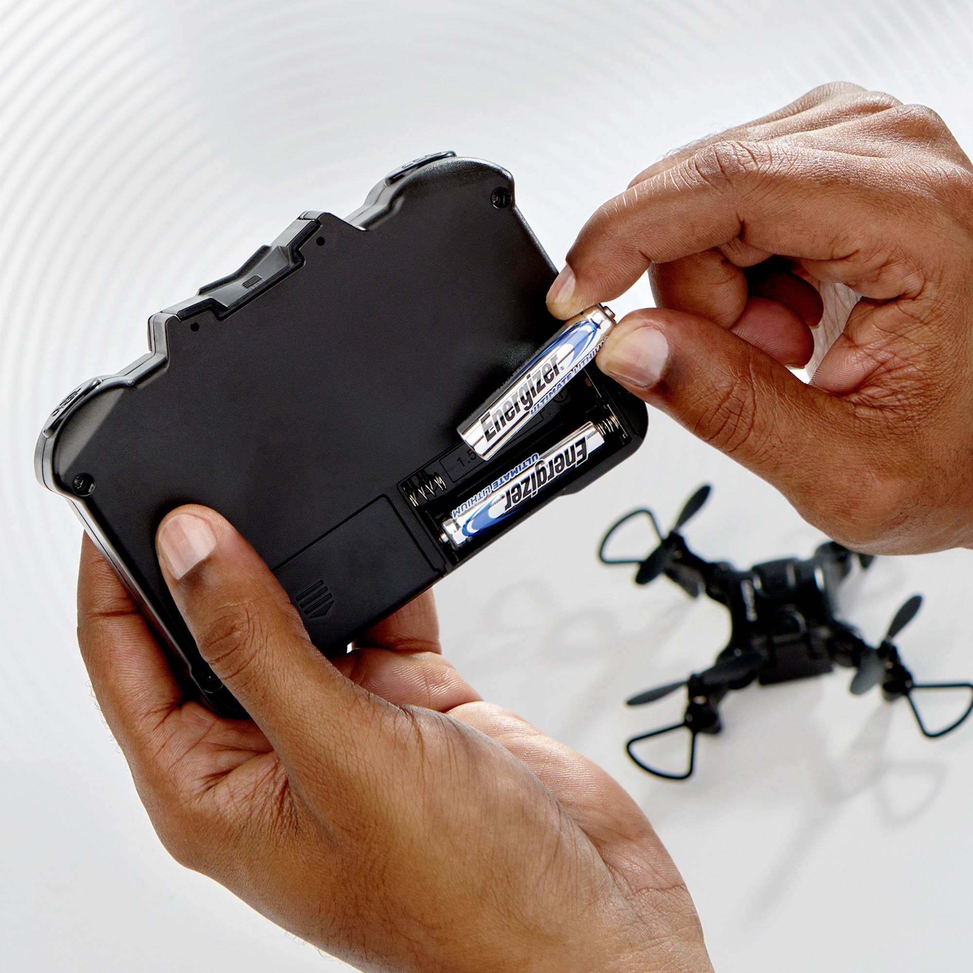 A person is changing batteries in a black remote control for a toy drone, which is lying on a table in the background.