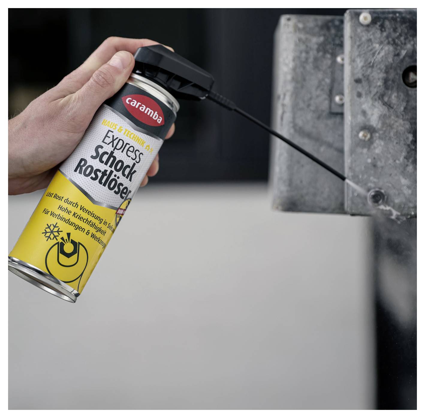 Hand sprays 'Caramba Express Schock-Rostlöser' on a stuck metal bolt or hinge to loosen rust on a gray mechanical part.