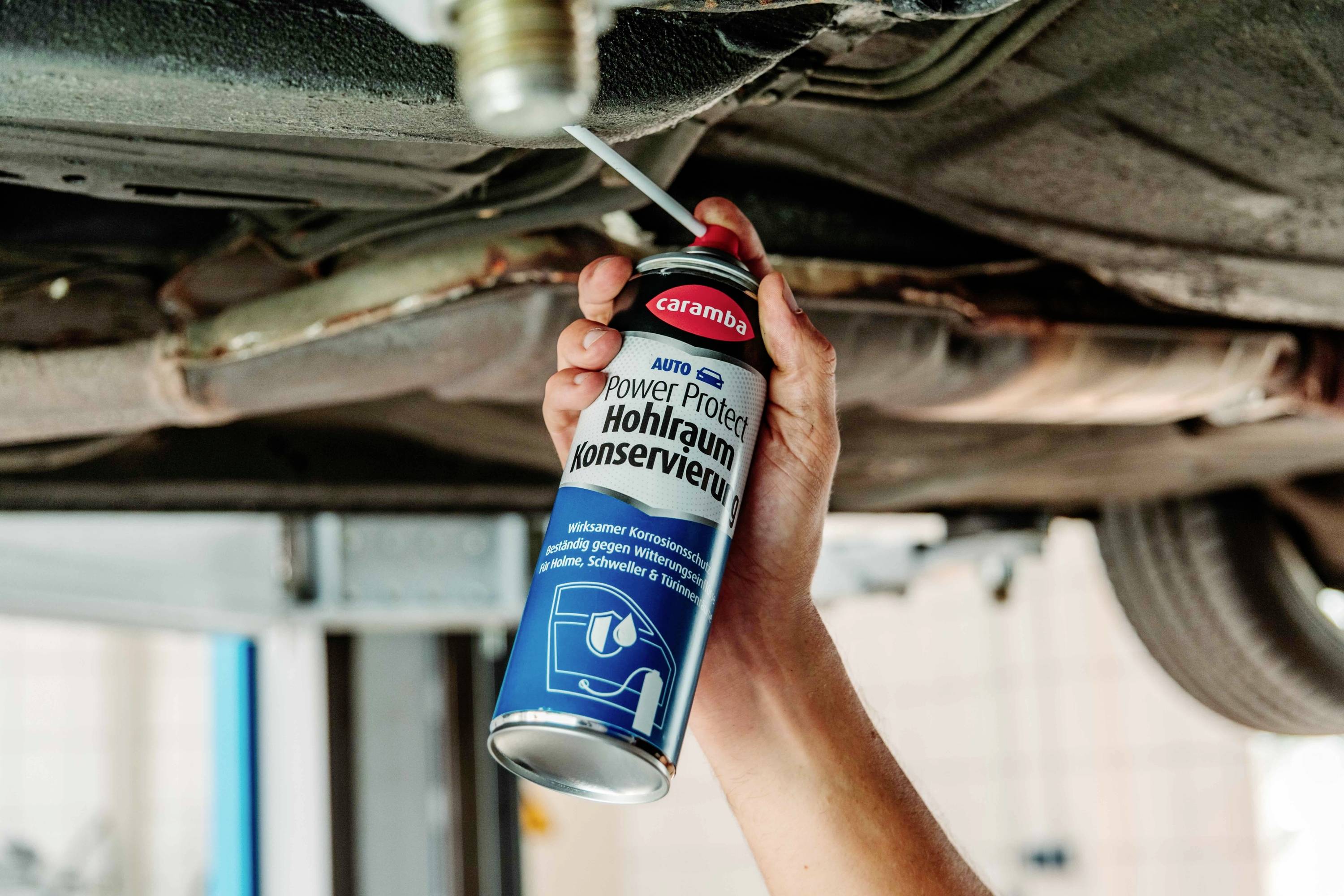 A person uses a spray can labeled 'Power Protect Hohlraum Konservieren' on the underside of a vehicle, likely applying rust protection.