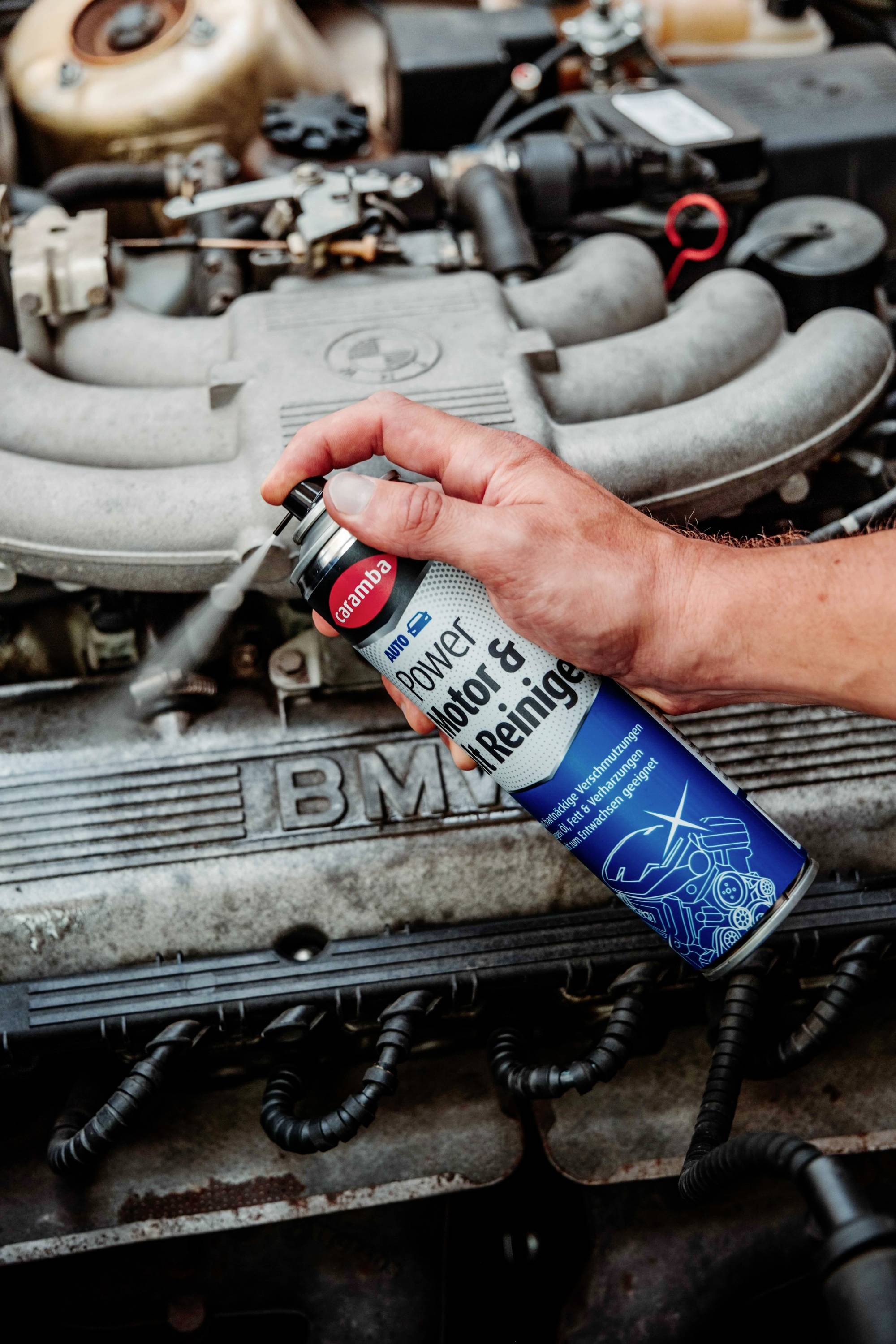 A person sprays cleaning fluid onto a car engine, focusing on the mechanical parts. The cleaner can has text and diagrams related to engine maintenance.