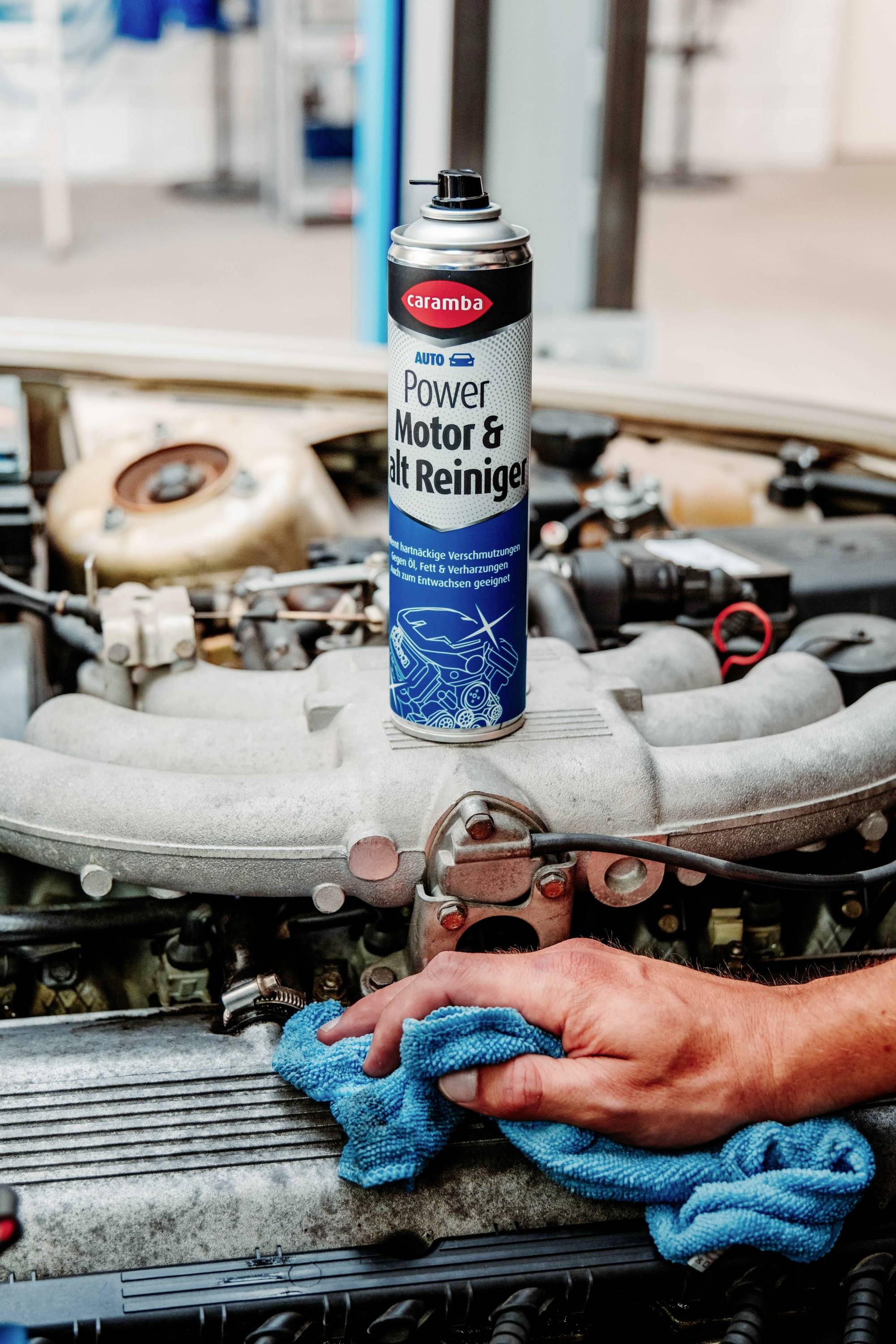A can of 'caramba Power Motor & Belt Reiniger' is on a car engine while a person uses a blue cloth to clean engine components.