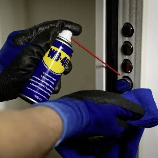 A pair of hands wearing blue gloves spraying WD-40 onto door hinges for lubrication to reduce noise.