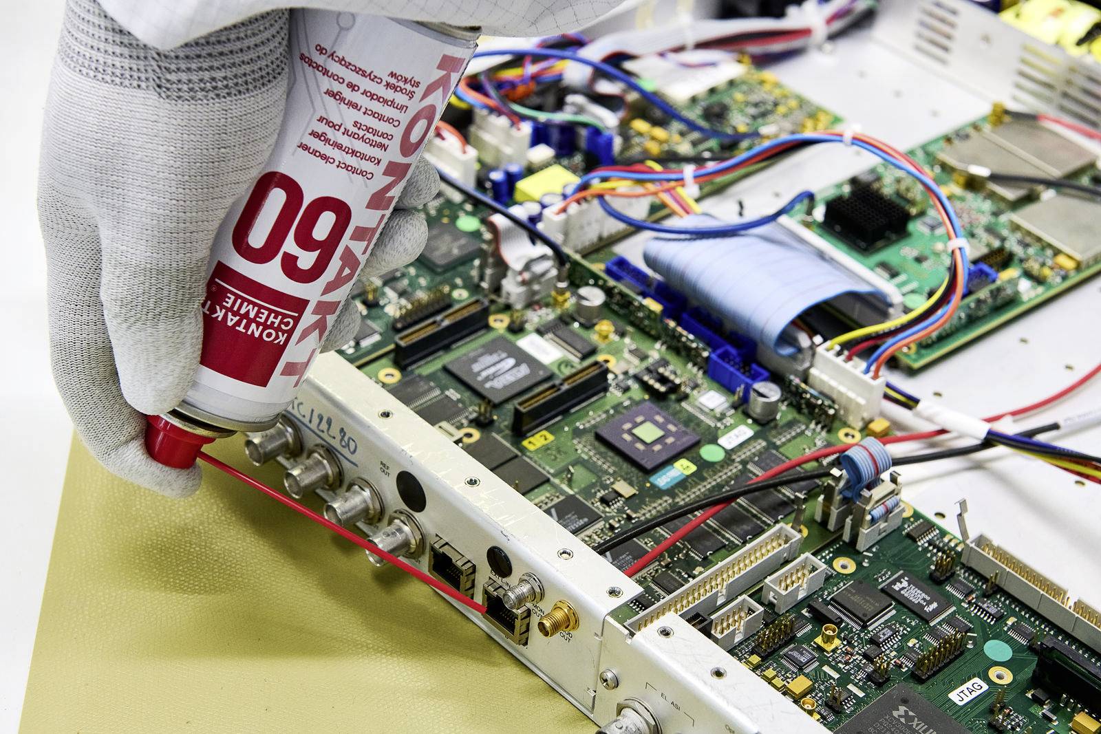 Worker wearing a glove applying thermal paste to an electronic circuit board. Various circuits and cables visible.