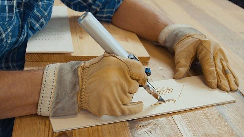 A person wearing gloves is engraving a pattern onto a wooden board with a specialised tool at a wooden table.