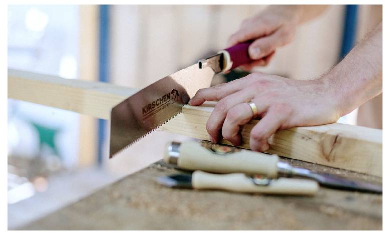 Close-up of hands using a hand saw to cut a wooden board on a workbench, with tools in the foreground.