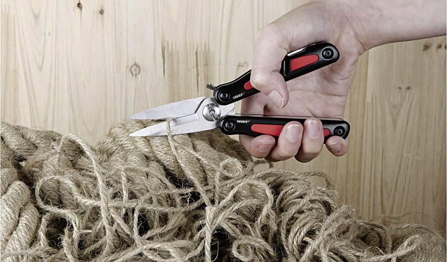 A hand holds a multi-functional tool with scissors, cutting through a bundle of coarse ropes on a wooden background.