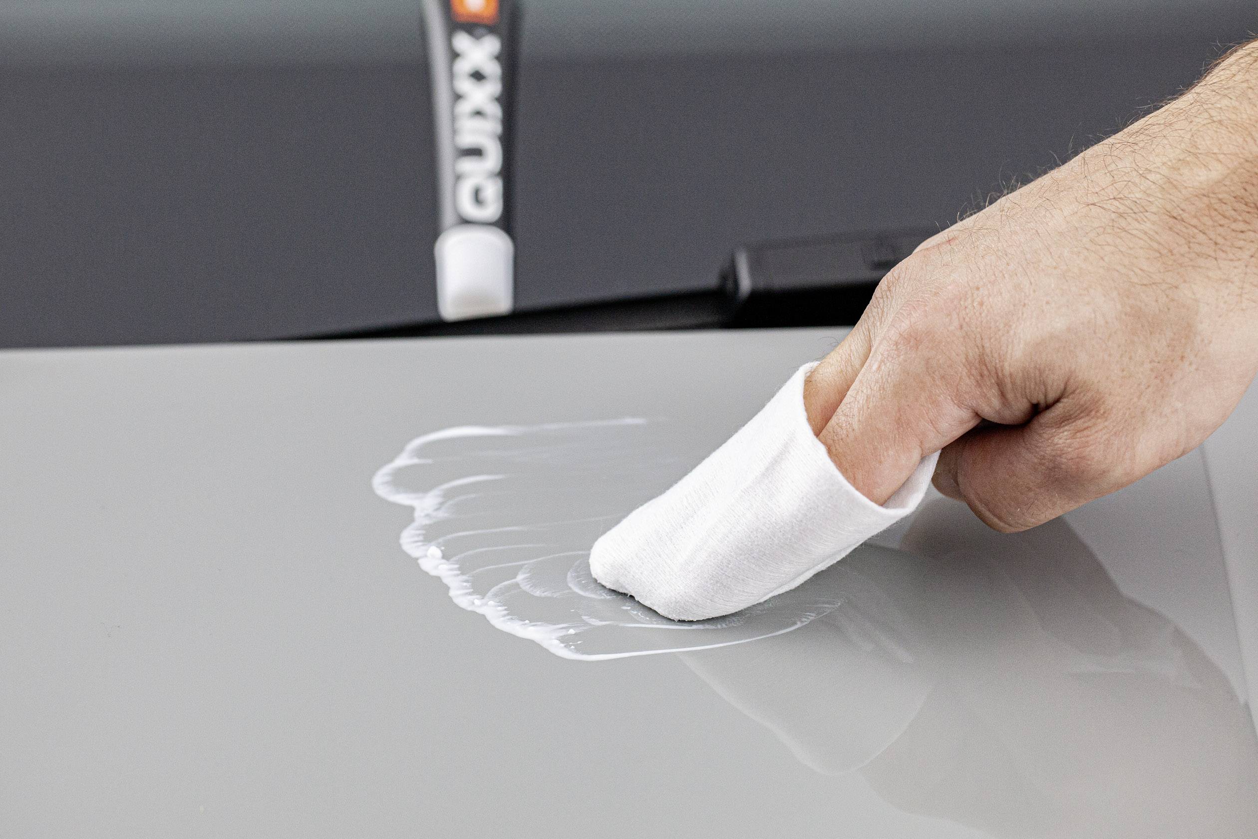 A hand with a white cloth polishes white paste onto grey car paintwork. In the background, a polishing compound tube is standing.
