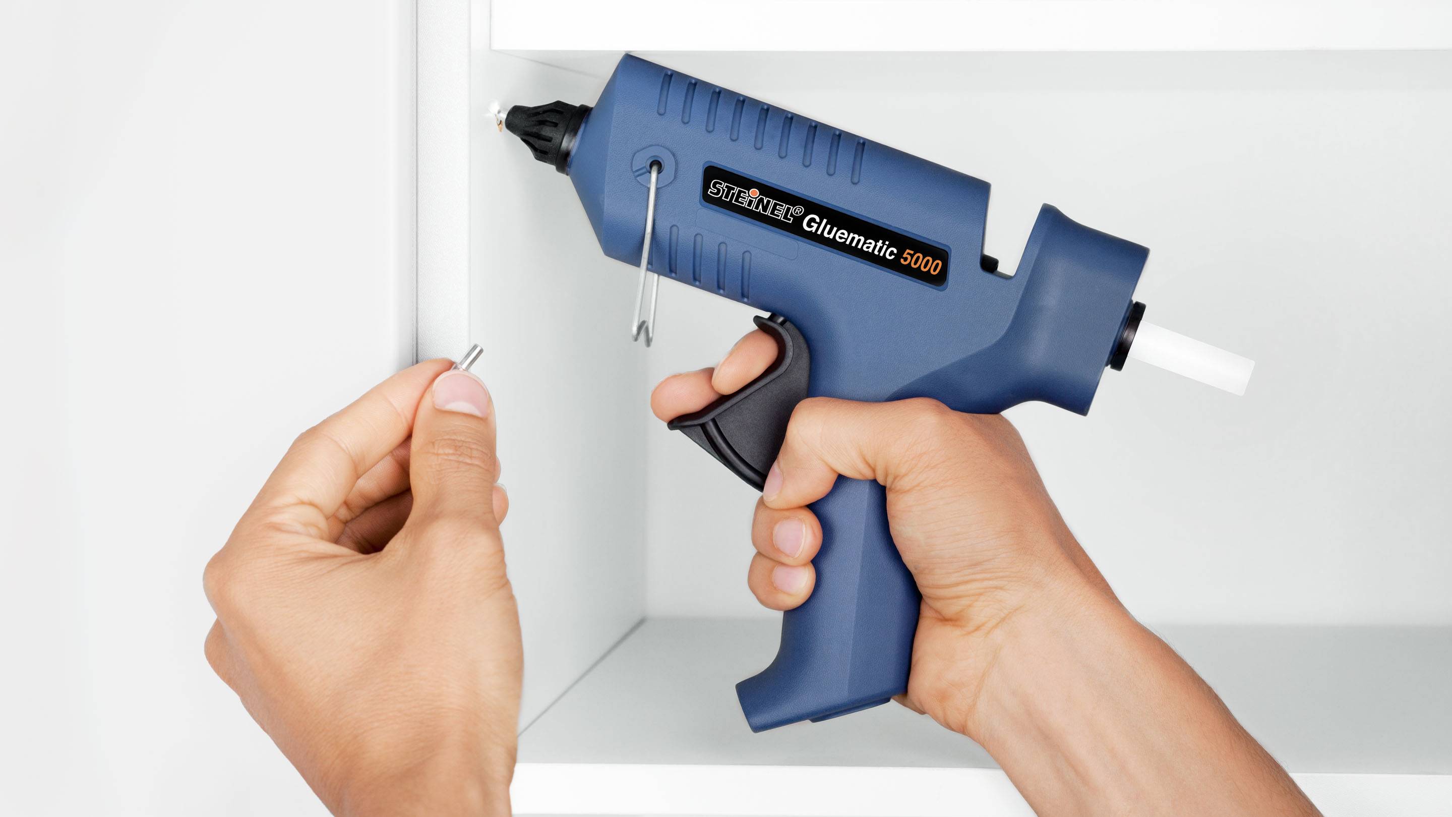 A person is holding a blue hot glue gun and a small nail in front of a white shelf. The hot glue gun bears the imprint 