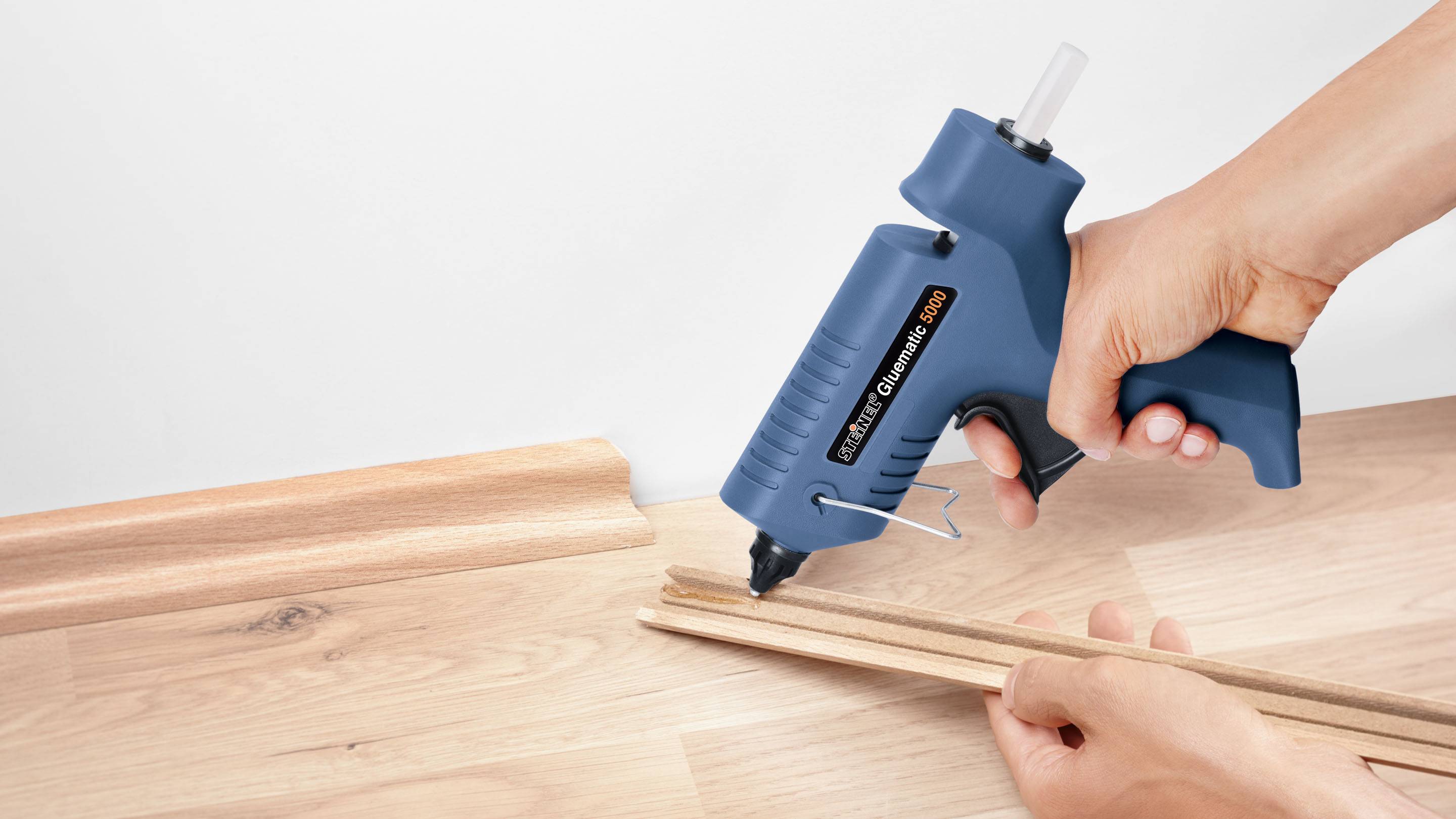A person is holding a blue hot glue gun and joining two wooden battens on a wooden floor.