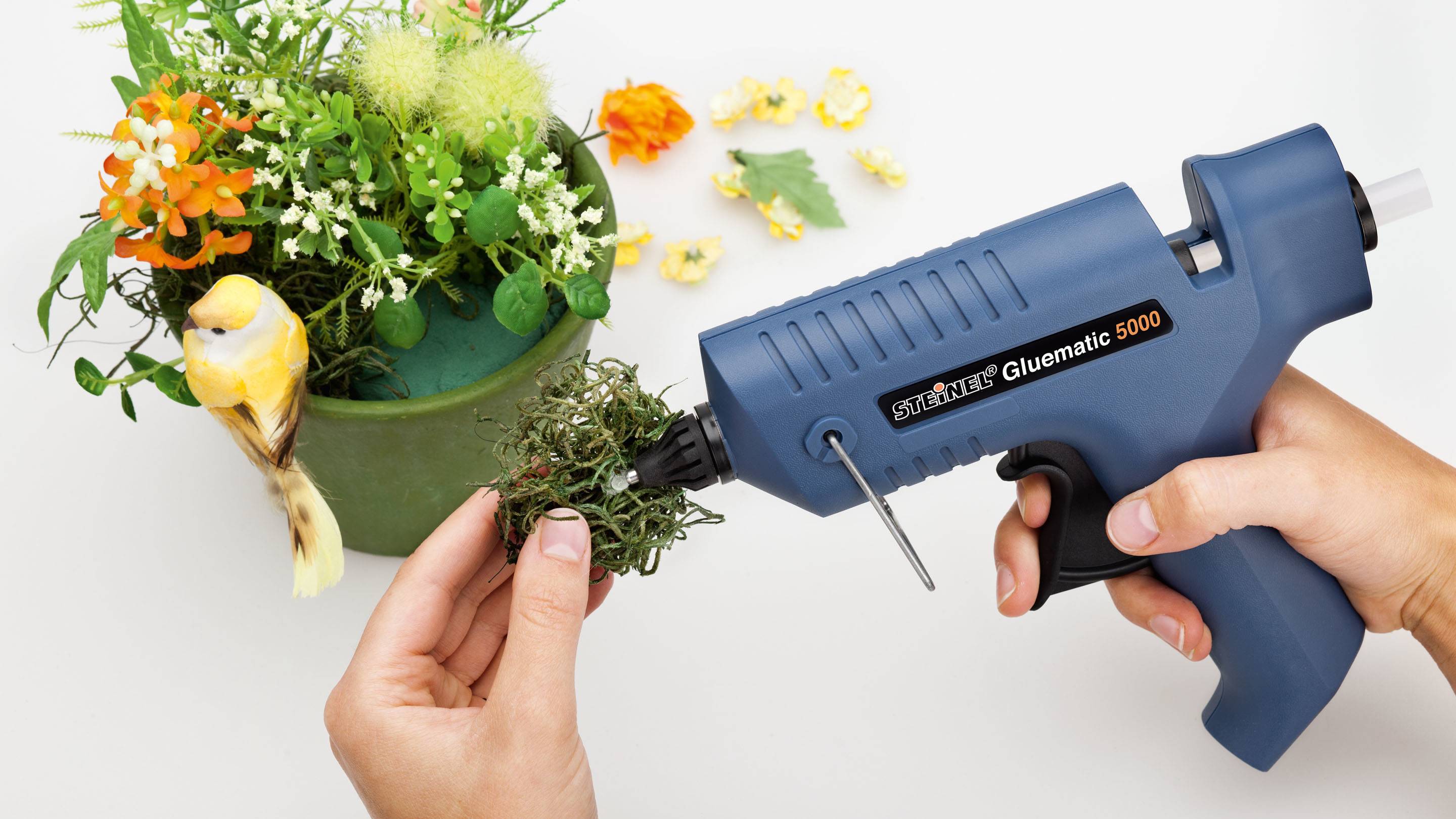 A person is decorating a green plant pot with artificial moss and a plastic hot glue gun. An artificial bird sits on the rim.