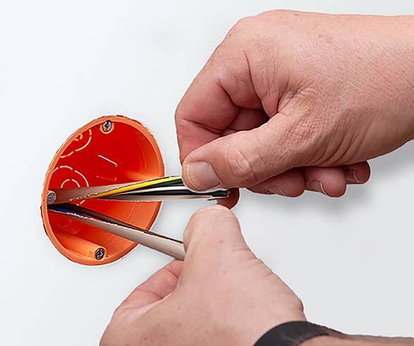 A person inserts electrical wires into an orange wall box, preparing for installation. Hands hold the wires and box securely.