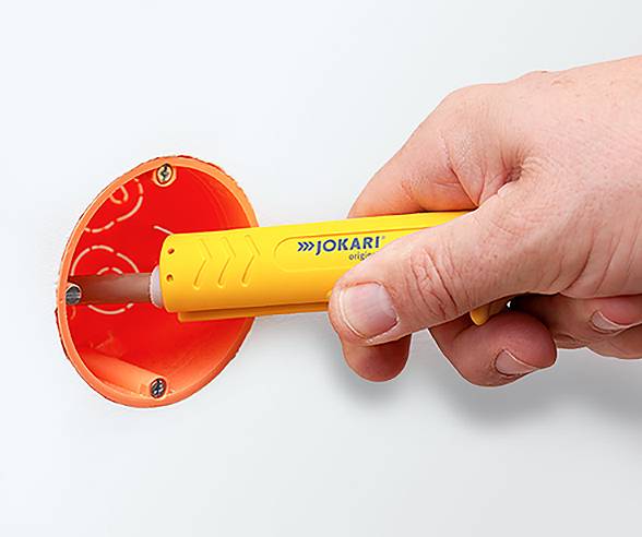 A hand is using a yellow cable stripping tool to prepare an orange electrical box in a wall for wiring installation.