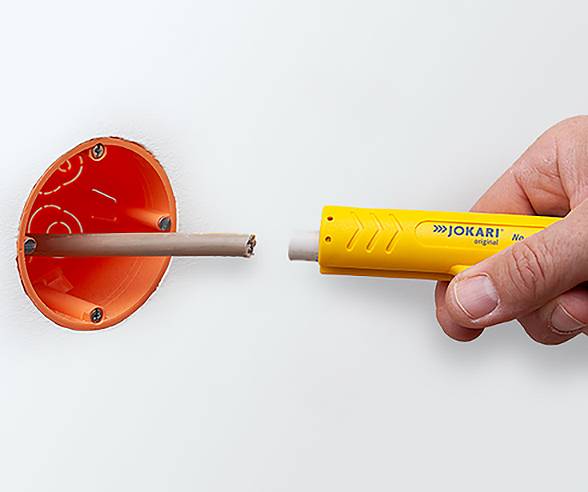 A hand uses a yellow wire stripping tool to strip insulation from a cable emerging from an orange wall socket opening.