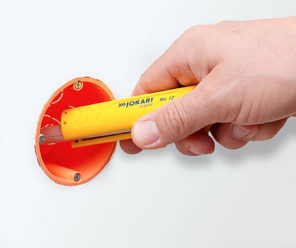A hand gripping a yellow cable stripping tool labeled 'Jokari' prepares to strip an orange wire in a wall.