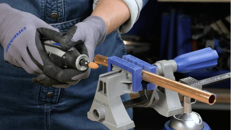 A person uses a rotary tool to polish a copper pipe held in a vise on a workbench. They are wearing gloves and a denim shirt.