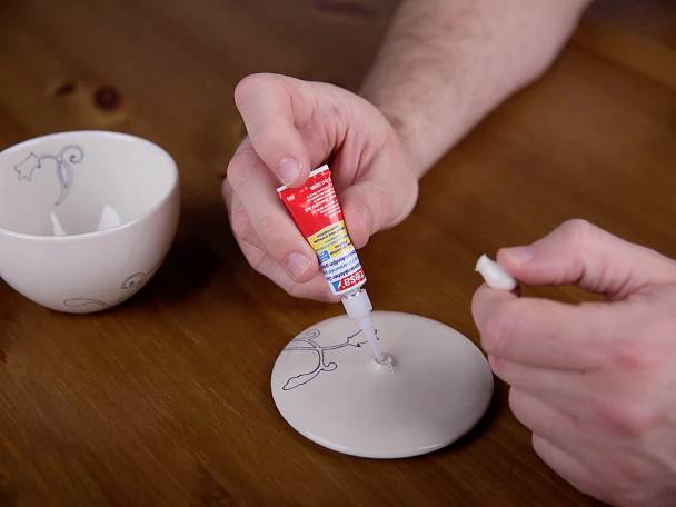 A person applies adhesive to mend a broken ceramic lid with hand-drawn designs, on a wooden table, with another broken piece nearby.