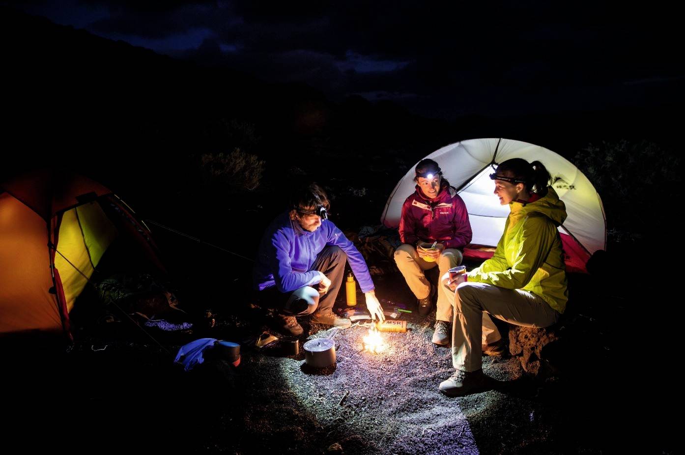 Drei Personen sitzen in einem dunklen Wald um ein Lagerfeuer vor zwei Zelten. Sie tragen Outdoor-Kleidung und genießen die Nacht.