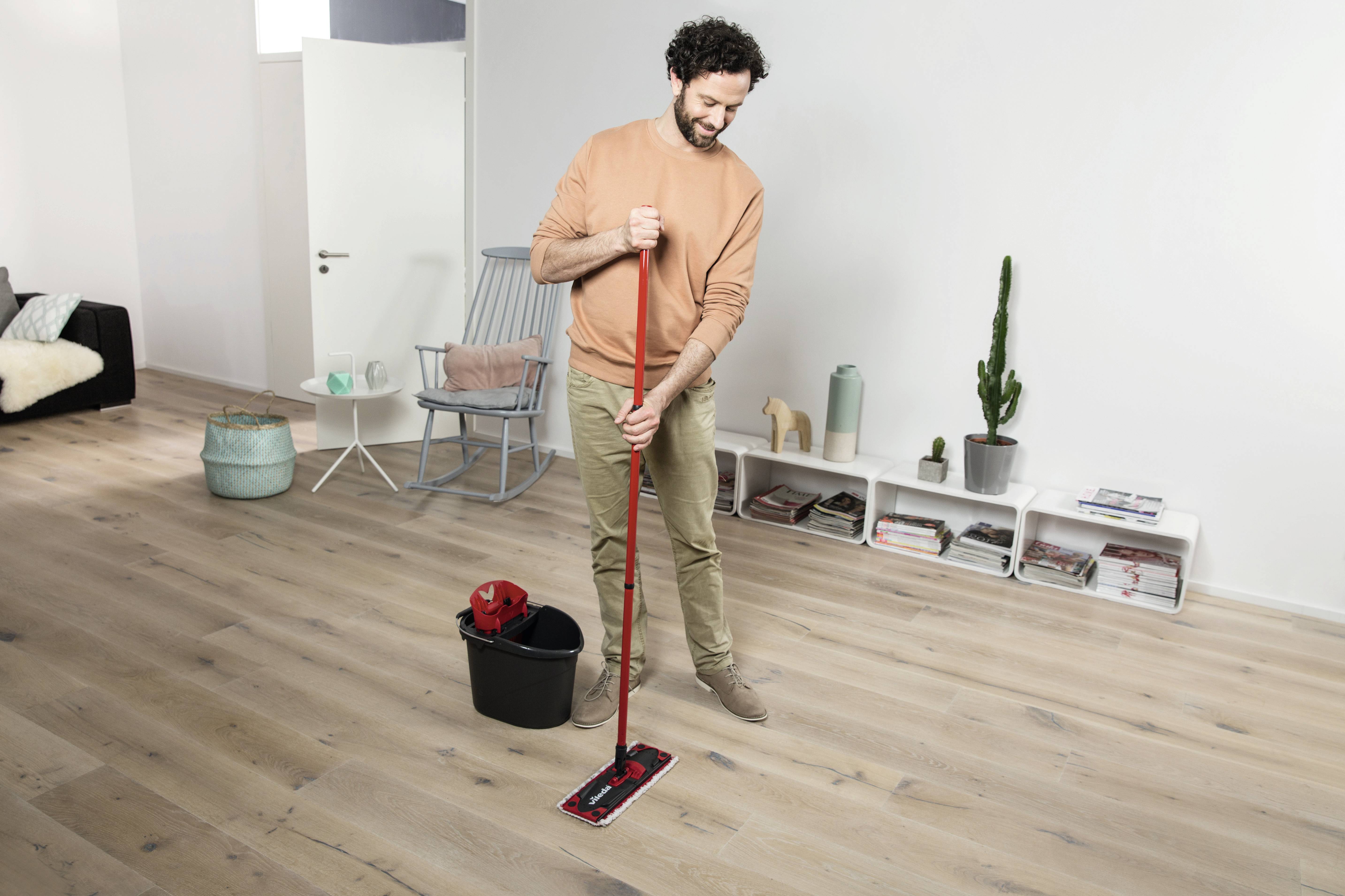 A man is mopping the wooden floor in a modern living room with a red mop; bucket and furniture in the background.