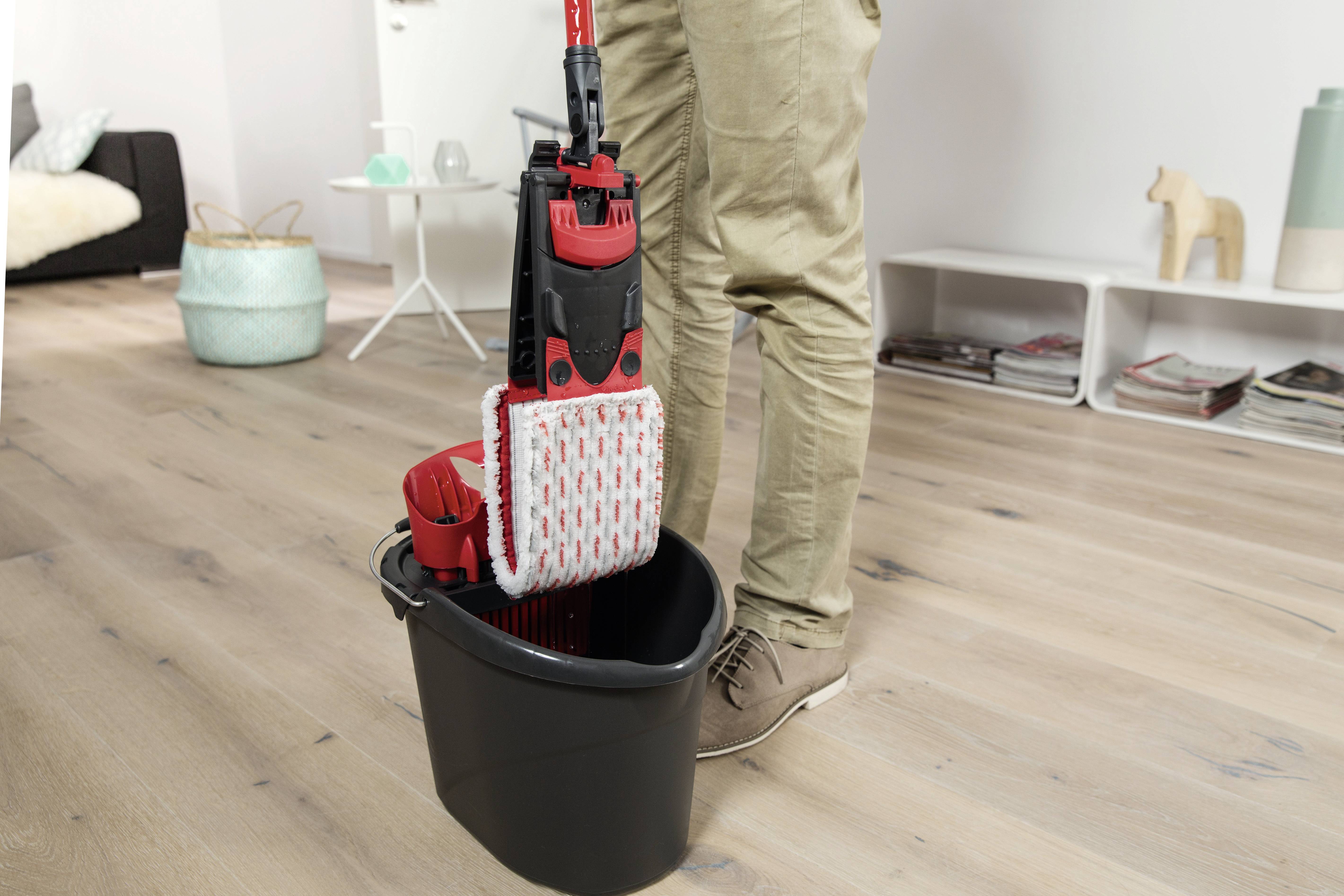A person is cleaning the floor with a mop and bucket in a living room. A sofa and shelving unit are visible in the background.