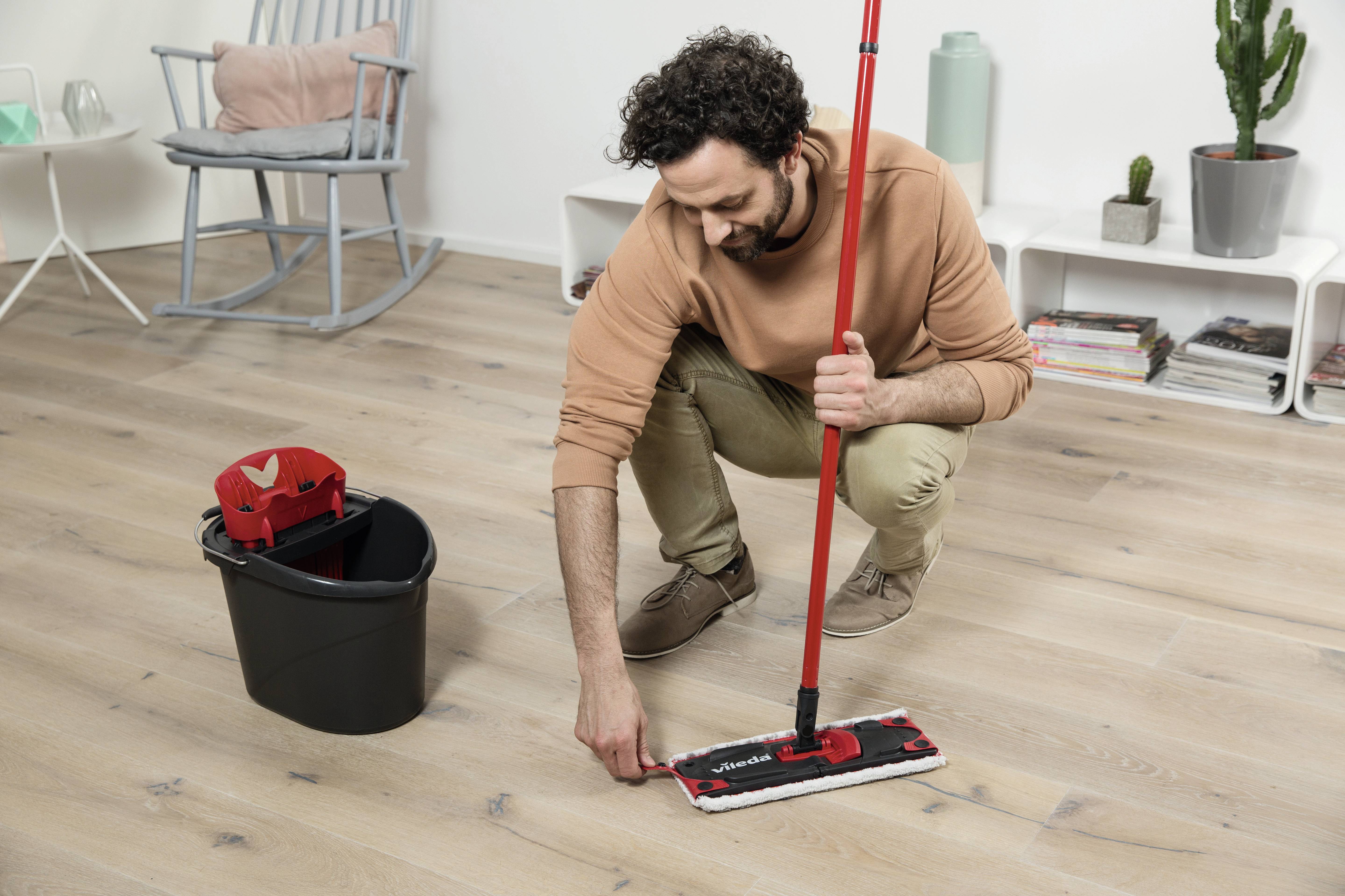 A man is mopping a wooden floor with a mop. A bucket stands beside him. In the background, an armchair and a bookshelf can be seen.
