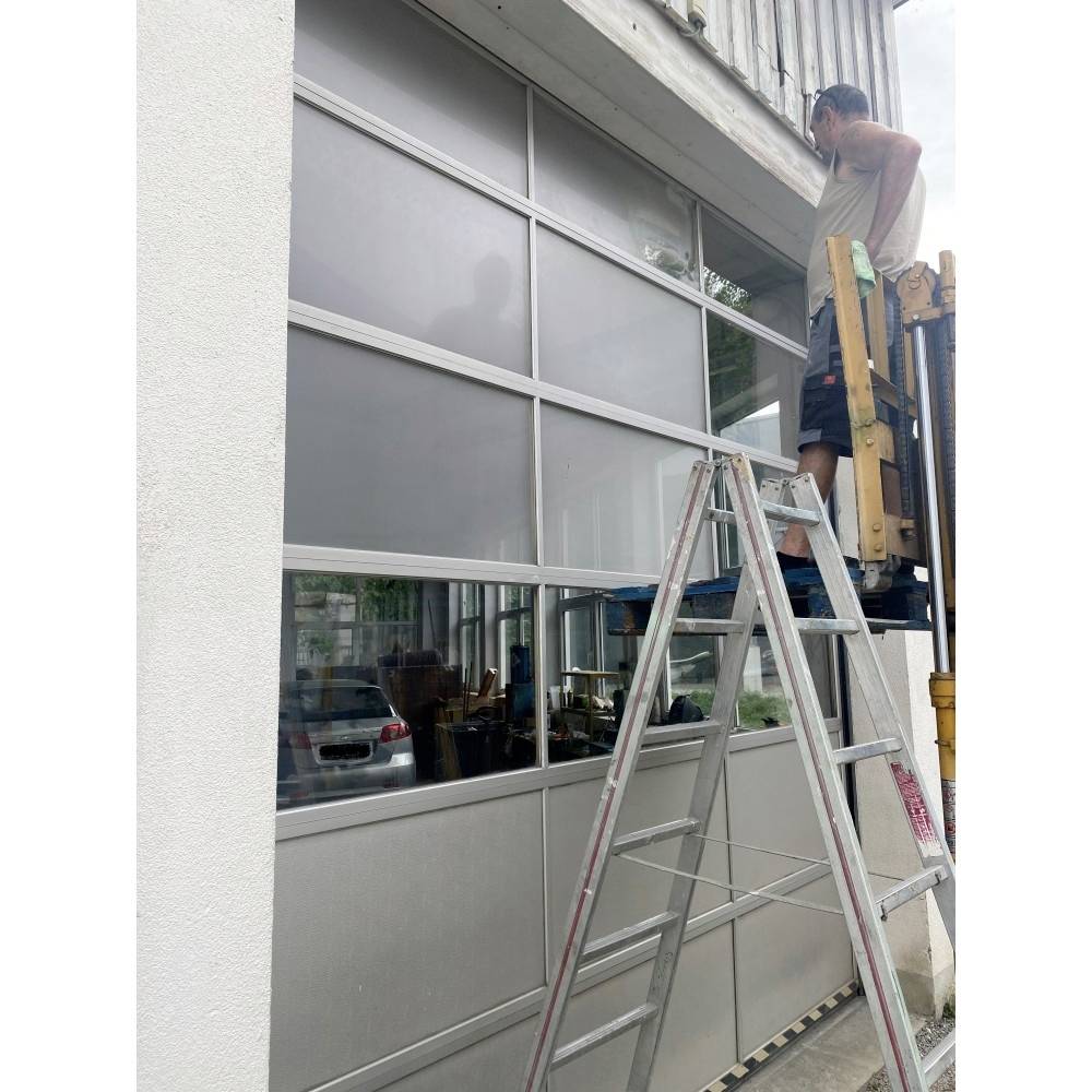 A man works on a garage door from a lifting platform, with a ladder nearby. The scene suggests maintenance or repair work.
