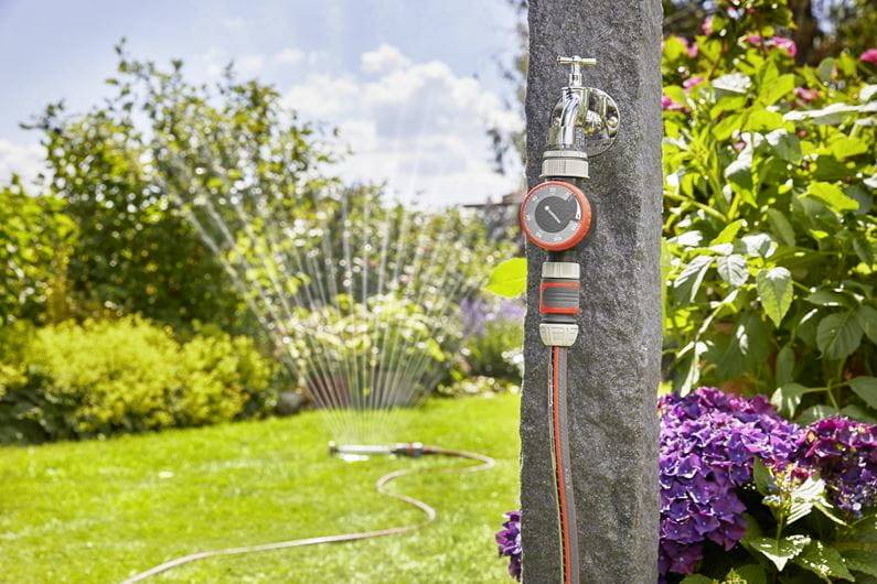A watering timer on a tap automatically controls garden irrigation. In the background, a lawn sprinkler is spraying water.