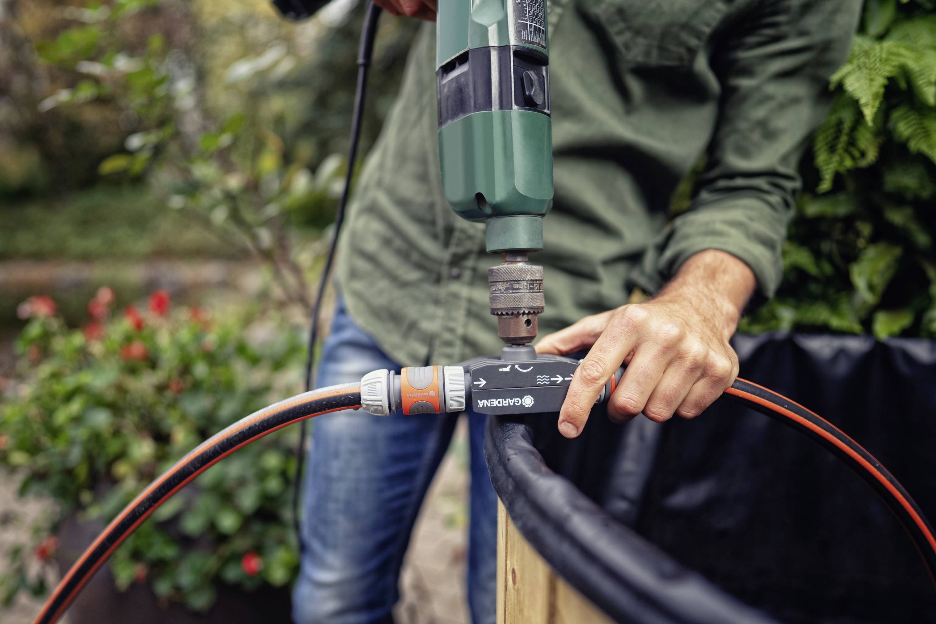 A person wearing a green shirt is using a drill to secure a connection on a garden hose. Plants are visible in the background.