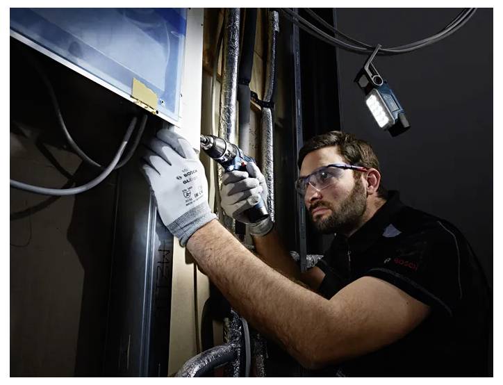 A man wearing safety glasses and gloves uses a power drill to work on a metal surface. He is focused and surrounded by pipes.