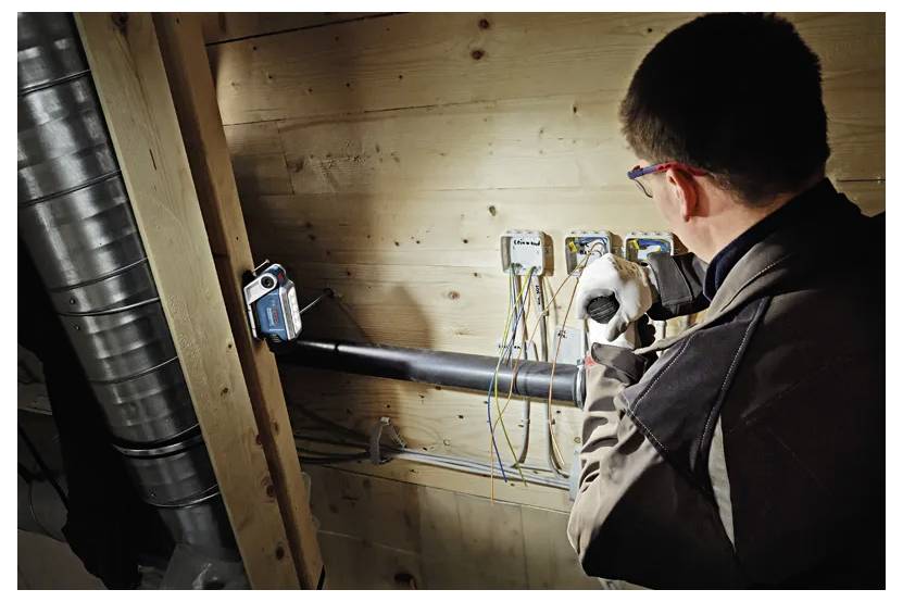 A person is working on electrical wiring inside a wooden structure, surrounded by conduits and insulated pipes. Safety gear is worn.